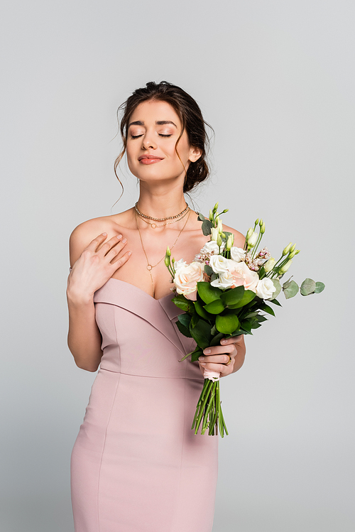 smiling woman holding wedding bouquet while standing with closed eyes isolated on grey