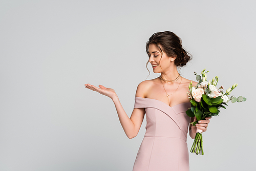 cheerful woman pointing with hand while standing with wedding bouquet isolated on grey