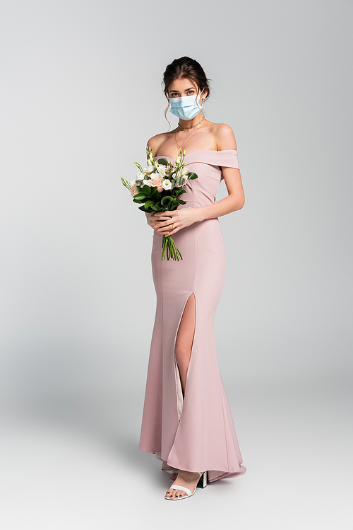 full length view of bride in medical mask posing with wedding bouquet on grey
