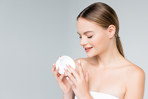 cheerful young woman holding container with face cream isolated on grey