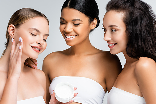 cheerful multiethnic women with bare shoulders looking at friend applying cosmetic cream isolated on grey