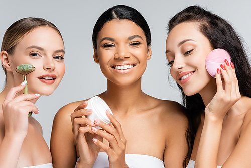 happy multiethnic women smiling while holding container with cosmetic cream, jade roller and silicone cleanser isolated on grey