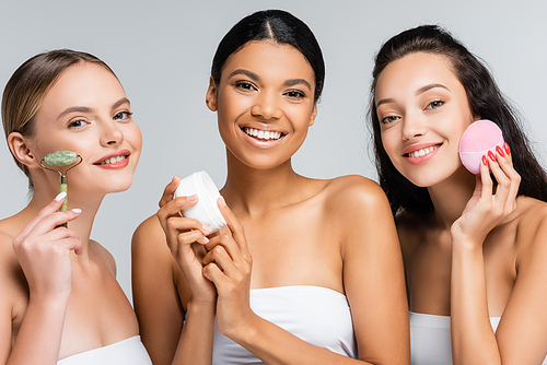 multiethnic women smiling while holding container with cosmetic cream, jade roller and silicone cleanser isolated on grey