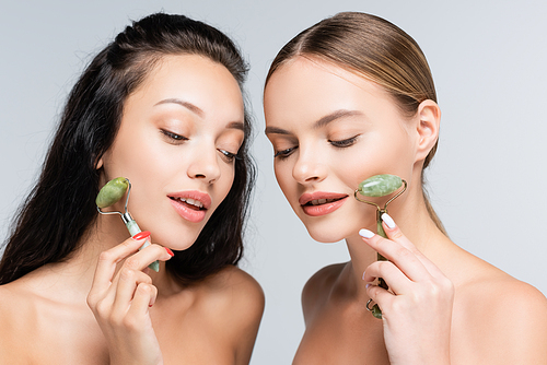 young women with bare shoulders smiling while holding jade rollers isolated on grey