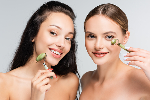 happy women with bare shoulders smiling while holding jade rollers isolated on grey