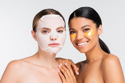 woman with moisturizing sheet mask near happy african american friend in golden eye patches isolated on white