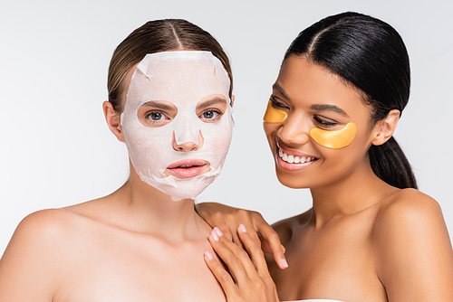young woman with moisturizing sheet mask near joyful african american friend in eye patches isolated on white