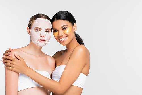 woman with moisturizing sheet mask near joyful african american friend in eye patches isolated on white