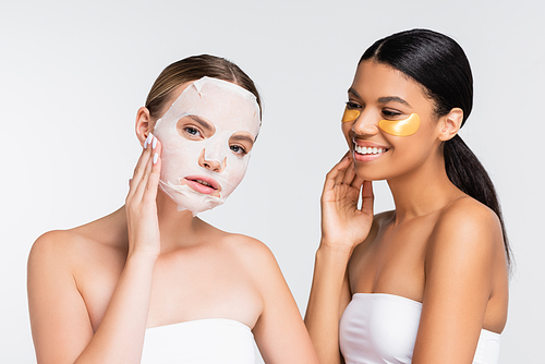 young woman applying moisturizing sheet mask near african american friend in eye patches isolated on white