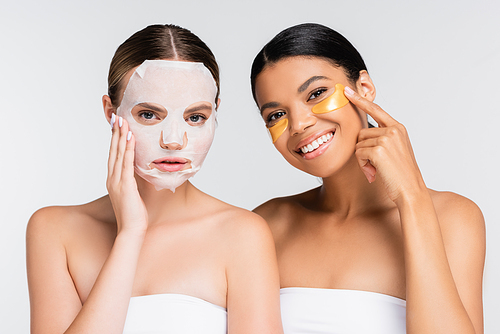 young woman with moisturizing sheet mask near cheerful african american friend in eye patches isolated on white