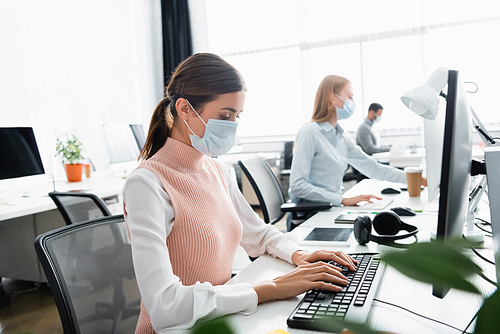 Businesswoman in medical mask using computer near colleagues on blurred background
