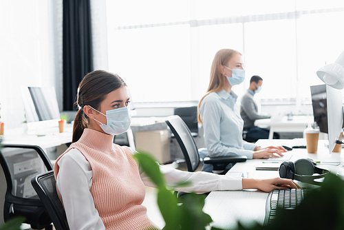 Businesswoman in medical mask sitting near computer keyboard and colleagues on blurred background