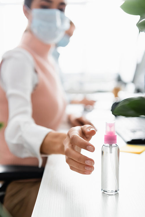 Businesswoman in medical mask pulling hand to sanitizer on table on blurred background