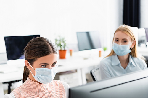 Businesswoman in medical mask looking at computer monitor near colleague in office
