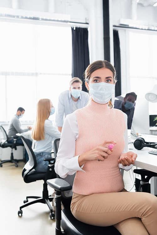 Businesswoman in medical mask using hand sanitizer while colleagues working on blurred background