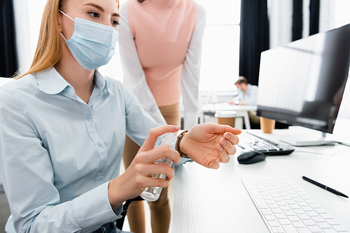 Businesswoman in medical mask using hand sanitizer near colleague and computer on blurred background