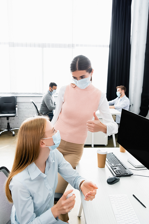 Businesswoman in medical mask showing like near colleague using hand sanitizer in office