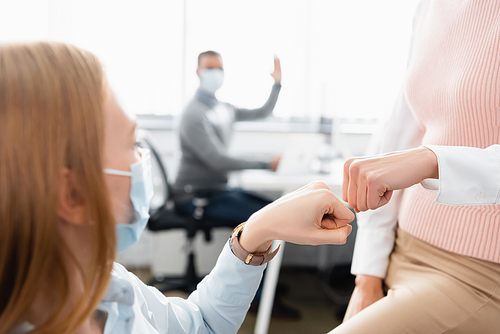 Businesswoman in medical mask doing fist bump with colleague on blurred background