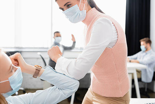 Businesswomen in medical masks giving high five with colleagues on blurred background in office