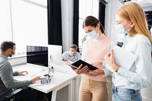 Businesswomen in medical masks with digital table looking at notebook near colleagues on blurred background