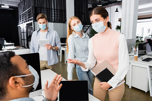 Businesswoman in medical mask with notebooks pointing at indian colleague on blurred background in office