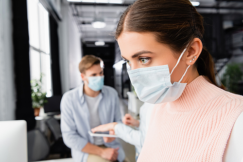 Businesswoman in medical mask standing near colleagues on blurred background in office