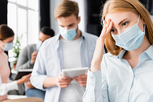 Sick businesswoman in medical mask touching head near colleagues on blurred background in office