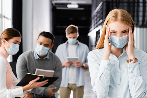 Diseased businesswoman in medical mask touching near while multiethnic colleagues working at blurred background