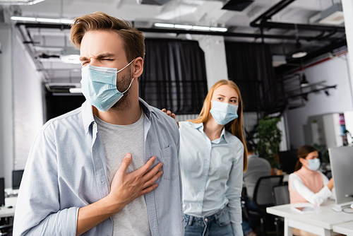 Diseased businessman in medical mask touching chest near colleague on blurred background in office
