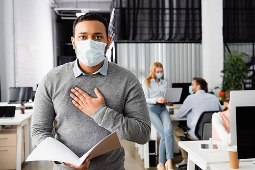 Indian businessman in medical mask touching chest while holding paper folder in office