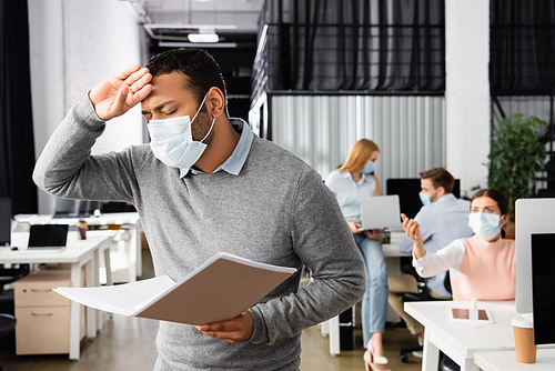Sick indian businessman in medical mask holding paper folder near colleagues on blurred background in office
