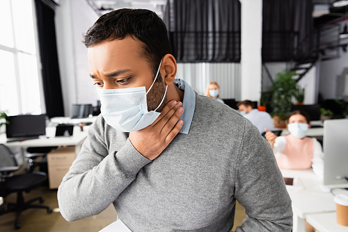 Diseased indian businessman in medical mask touching neck in office