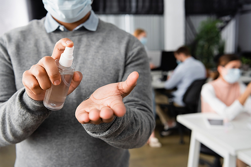 Cropped view of businessman in medical mask holding bottle of hand sanitizer in office