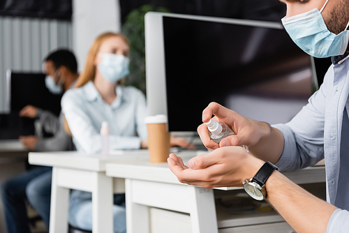 Cropped view of businessman in medical mask using hand sanitizer near computers and colleagues on blurred background