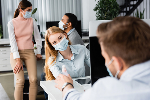 Businesswoman in medical mask giving hand sanitizer to colleague on blurred foreground in office