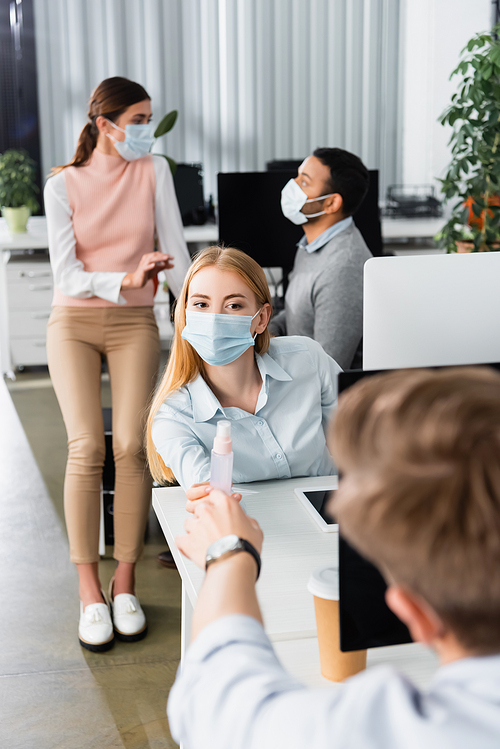 Businesswoman in medical mask giving hand sanitizer to colleague on blurred foreground while working in office