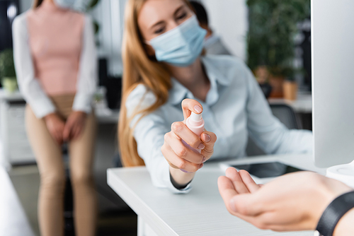 Businesswoman in medical mask spraying hand sanitizer on hand of colleague on blurred foreground in office