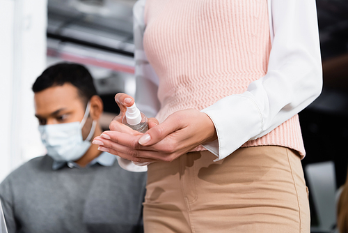 Cropped view of businesswoman using hand sanitizer near indian colleague in medical mask on blurred background