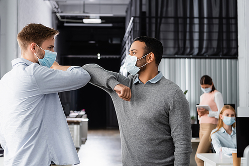 Multiethnic businessmen in medical mask giving high five with elbows near colleagues on blurred background in office