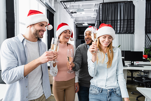 Positive multiethnic businesspeople in santa hats holding glasses of champagne in office