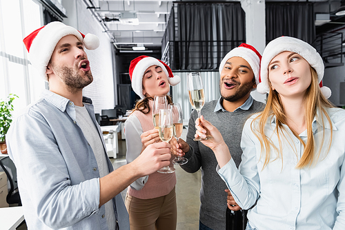 Excited multicultural businesspeople in santa hats holding glasses of champagne in office