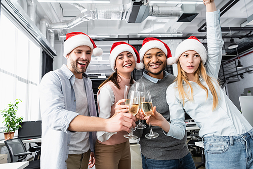 Positive multiethnic businesspeople in santa hats clinking with champagne in office