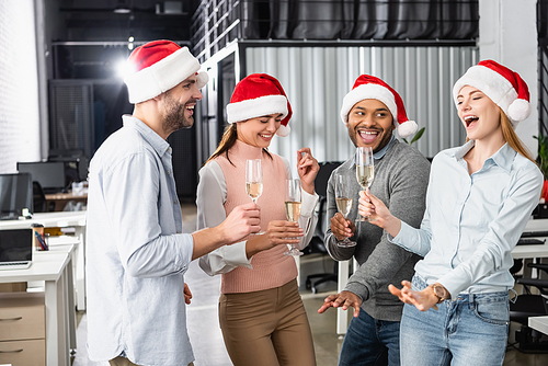 Cheerful multiethnic businesspeople in santa hats having fun with glasses of champagne in office