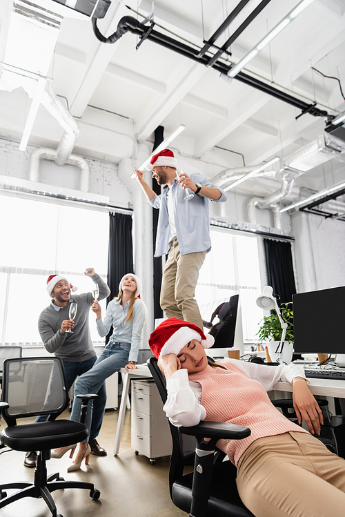 Businesswoman sleeping in santa hat near cheerful multiethnic colleagues with champagne on blurred background