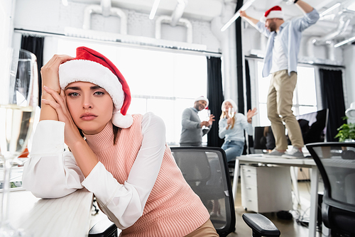 Tired businesswoman in santa hat  near glass of champagne and colleagues on blurred background in office
