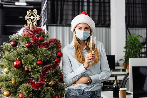 Businesswoman in medical mask and santa hat holding glass of champagne near christmas tree in office