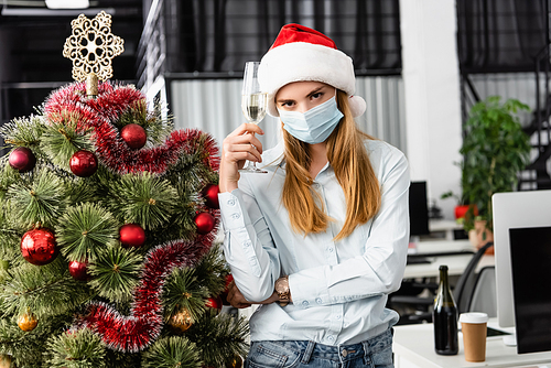 Businesswoman in medical mask holding glass of champagne near christmas tree in office