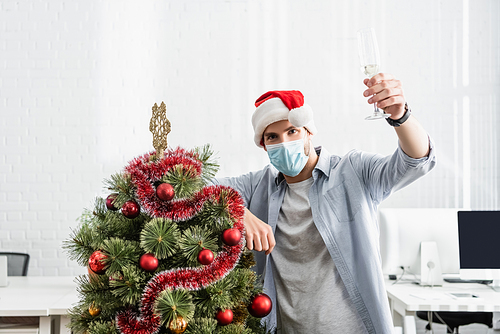 Businessman in medical mask and santa hat holding glass of champagne near christmas tree in office