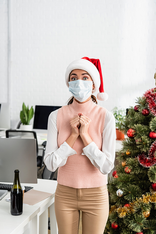 Young businesswoman in santa hat and medical mask standing near christmas tree and bottle of champagne in office