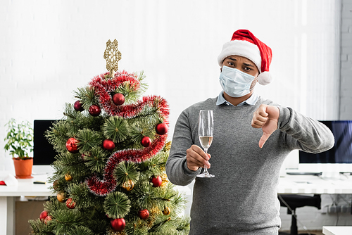 Indian businessman in medical mask holding glass with champagne and showing dislike gesture near christmas tree in office